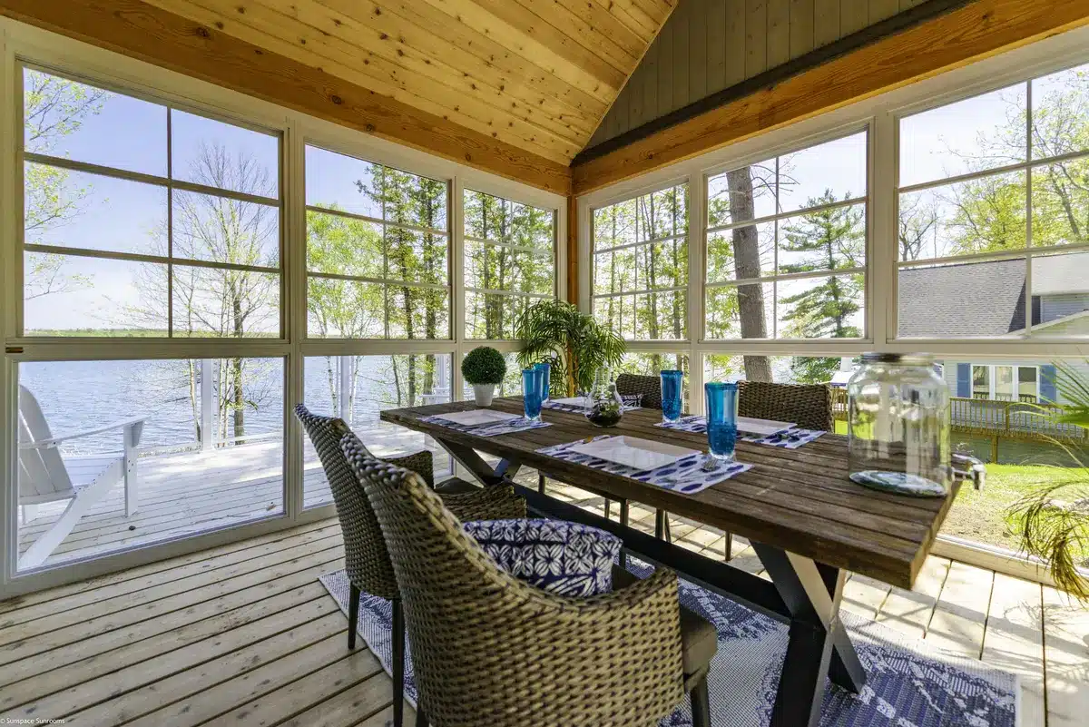 Interior of a sunroom with white frames and large WeatherMaster windows featuring a wooden dining table and lake view.